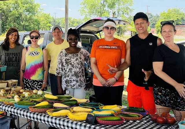 Leo and a group gathered behind a table of fresh vegetables at a community market.