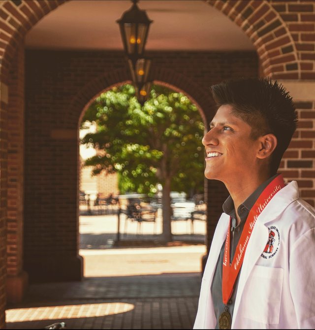 Leo in a white coat and graduation medallion beneath a campus archway.
