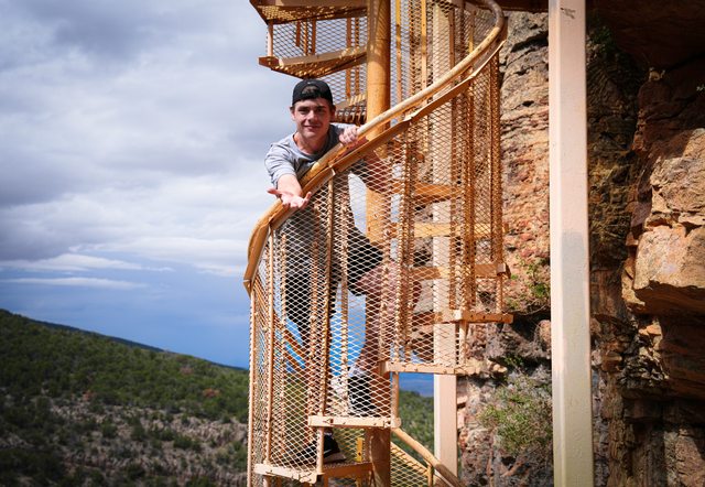 Chris on a golden spiral staircase beside a stone tower, with forest and hills visible beyond.