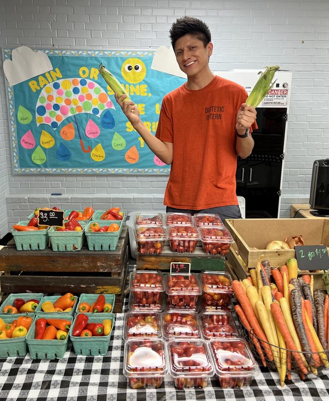 Leo holding fresh corn behind a community produce display.