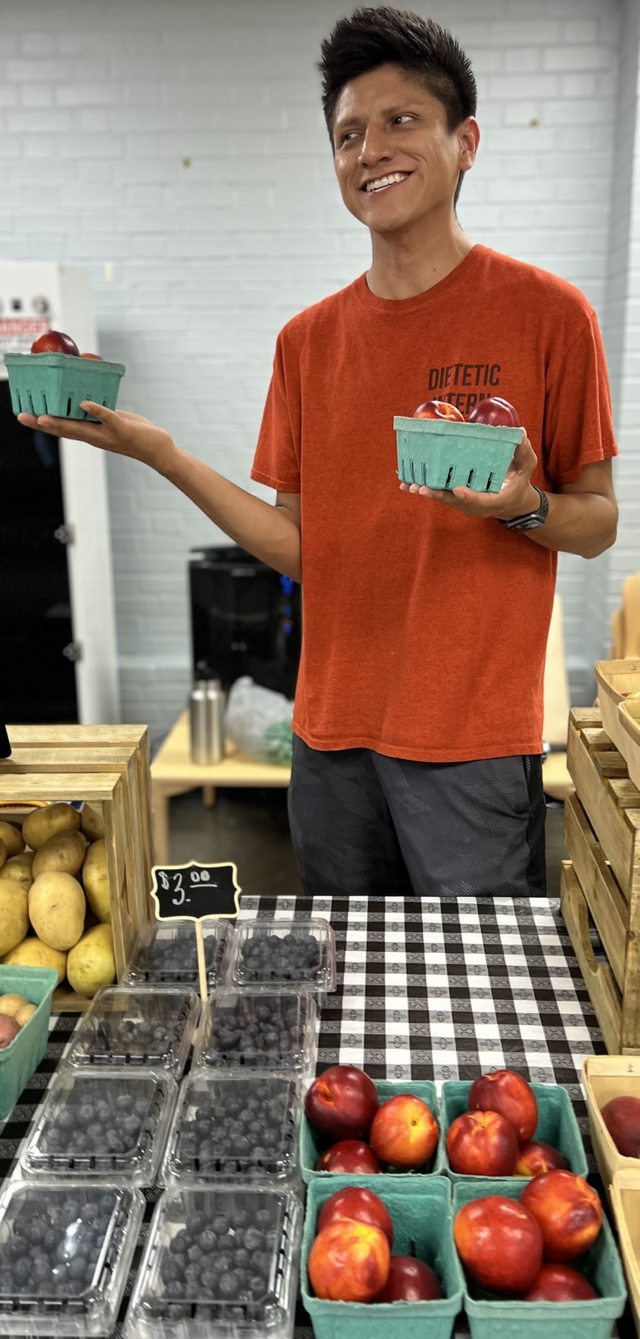 Leo holding baskets of fresh fruit beside a colorful community produce display.